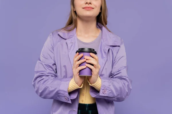 Vue recadrée de jeune femme souriante en trench coat tenant tasse en papier isolé sur violet — Photo de stock