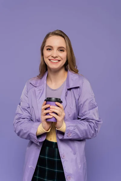 Cheerful young woman in trench coat holding paper cup isolated on purple — Stock Photo