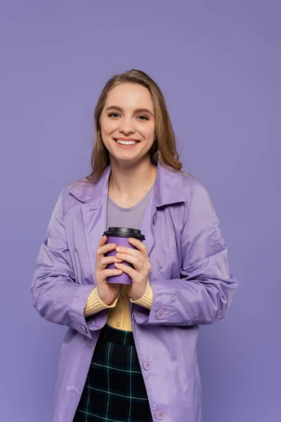 Allegra giovane donna in trench contenente tazza di carta isolata su viola — Foto stock