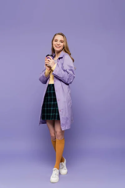 Full length of happy young woman in trench coat holding paper cup on purple — Stock Photo