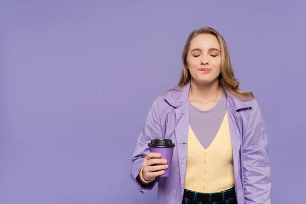 Pleased young woman in trench coat holding paper cup and biting lip isolated on purple — Stock Photo