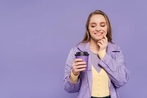 Heureuse jeune femme en trench coat regardant tasse en papier isolé sur violet — Photo de stock