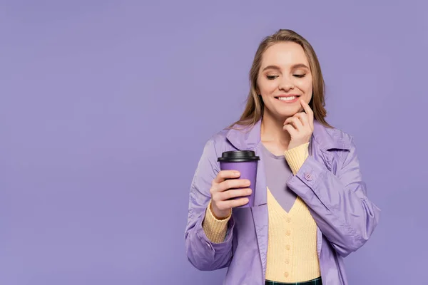 Felice giovane donna in trench guardando tazza di carta isolata su viola — Foto stock