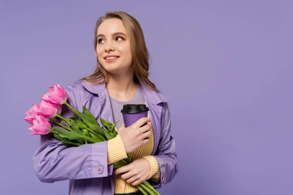 Happy young woman in trench coat holding paper cup and pink tulips isolated on purple — Stock Photo