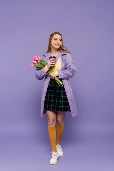 Pleine longueur de joyeuse jeune femme en trench coat tenant tasse en papier et tulipes roses sur violet — Photo de stock
