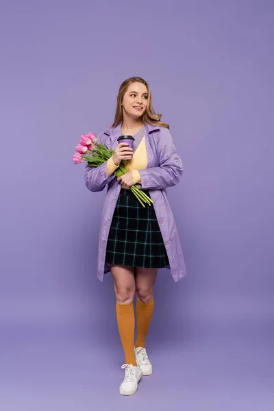 Comprimento total de alegre jovem mulher em casaco de trincheira segurando copo de papel e tulipas rosa em roxo — Fotografia de Stock