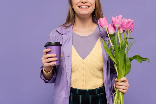 Cropped view of happy young woman in trench coat holding paper cup and pink tulips isolated on purple — Stock Photo
