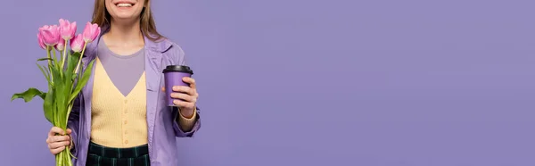 Vue recadrée d'une jeune femme heureuse en trench coat tenant une tasse en papier et des tulipes roses isolées sur violet, bannière — Photo de stock