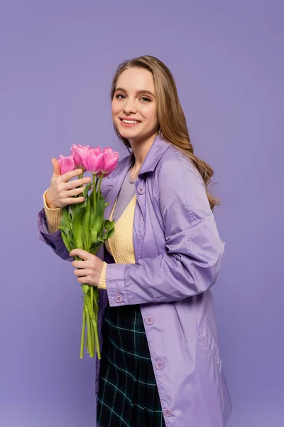 Feliz joven en gabardina sosteniendo tulipanes rosados aislados en púrpura - foto de stock