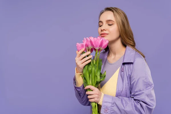 Young woman in trench coat smelling pink tulips isolated on purple — Stock Photo