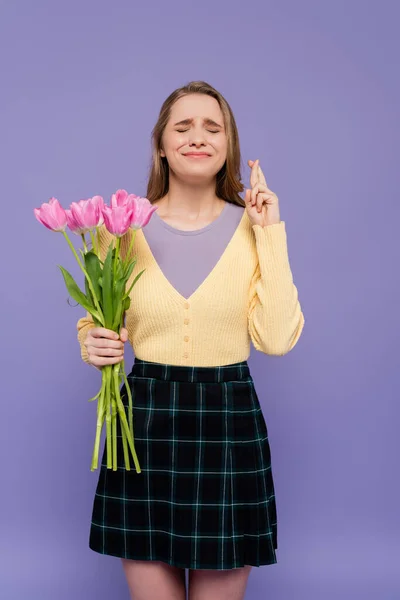 Jovem segurando tulipas rosa e de pé com os dedos cruzados isolados em roxo — Fotografia de Stock