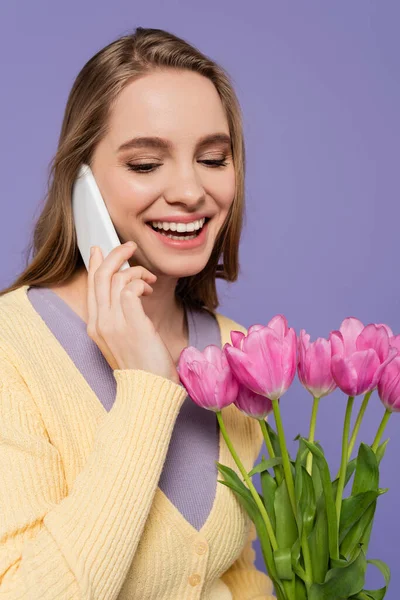 Веселая молодая женщина, держащая розовые тюльпаны и разговаривая на смартфоне изолированы на фиолетовый — стоковое фото