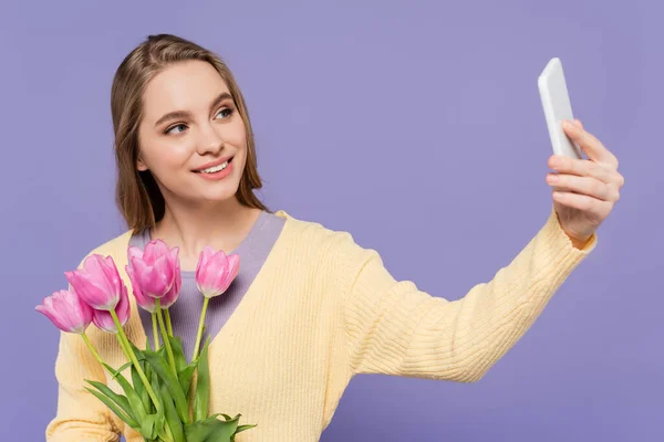 Allegra giovane donna che tiene tulipani rosa e prende selfie isolato su viola — Foto stock