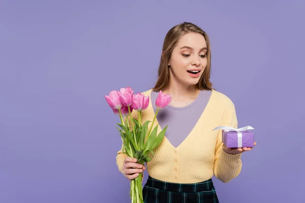Здивована молода жінка тримає рожеві тюльпани і подарункову коробку ізольовано на фіолетовому — стокове фото