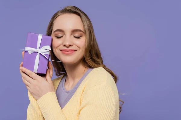 Mujer joven complacida sosteniendo envuelto caja de regalo aislado en púrpura - foto de stock