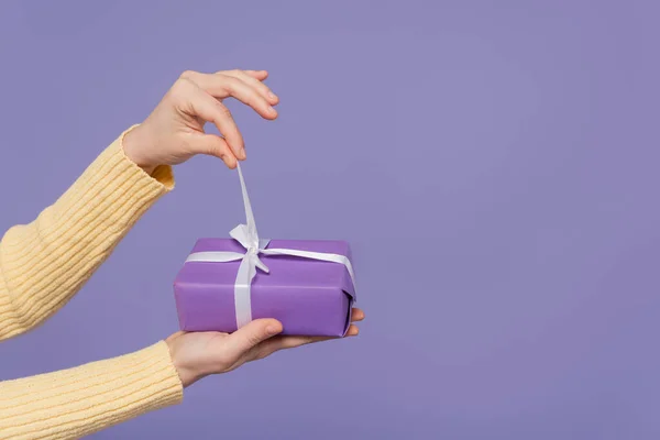 Vista recortada de la joven mujer tirando de la cinta en caja de regalo envuelto aislado en púrpura - foto de stock