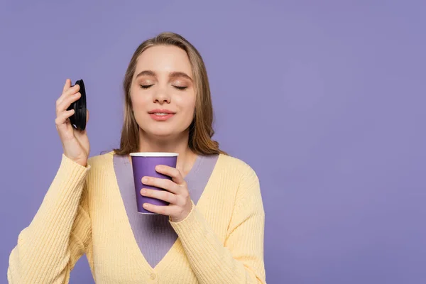 Jovem mulher cheirando café para ir em copo de papel isolado em roxo — Fotografia de Stock