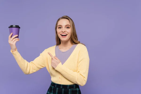 Mujer joven excitada señalando el café para ir en taza de papel aislado en púrpura - foto de stock