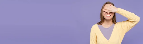 Mujer joven y feliz cubriendo los ojos aislados en púrpura, bandera - foto de stock