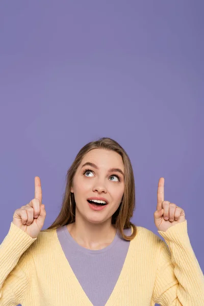 Изумленная молодая женщина смотрит вверх и указывает пальцами, изолированными на фиолетовый — Stock Photo