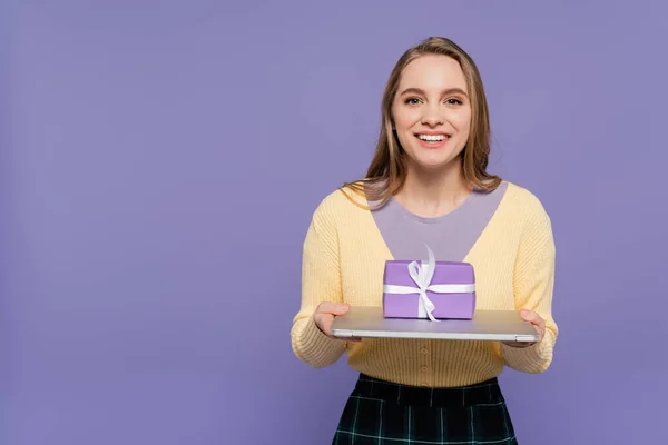 Glückliche junge Frau hält Laptop mit Geschenkbox isoliert auf lila — Stockfoto