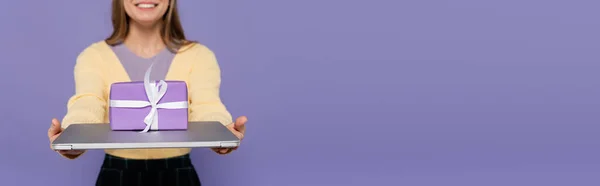Vue recadrée de la jeune femme heureuse tenant ordinateur portable avec boîte cadeau isolé sur violet, bannière — Photo de stock