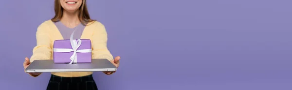 Cropped view of happy young woman holding laptop with gift box isolated on purple, banner — Stock Photo