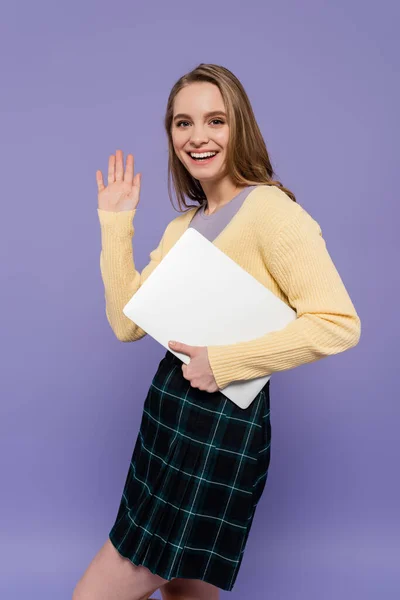 Fröhliche Studentin winkt mit der Hand und hält Laptop isoliert auf lila — Stockfoto