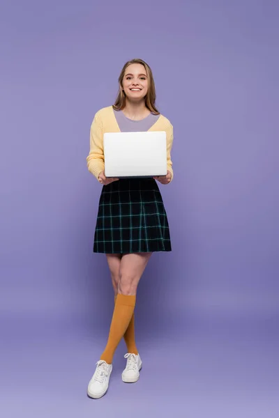 Longitud completa de alegre joven mujer sosteniendo el ordenador portátil en púrpura - foto de stock