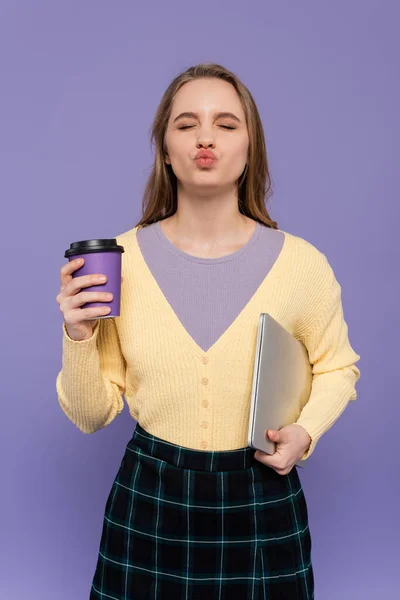 Молода жінка кидає губи, тримаючи ноутбук і паперову чашку ізольовано на фіолетовому — стокове фото