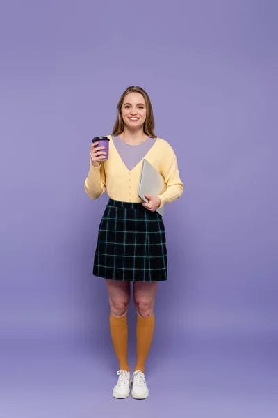 Comprimento total da jovem mulher feliz segurando laptop e copo de papel em roxo — Fotografia de Stock