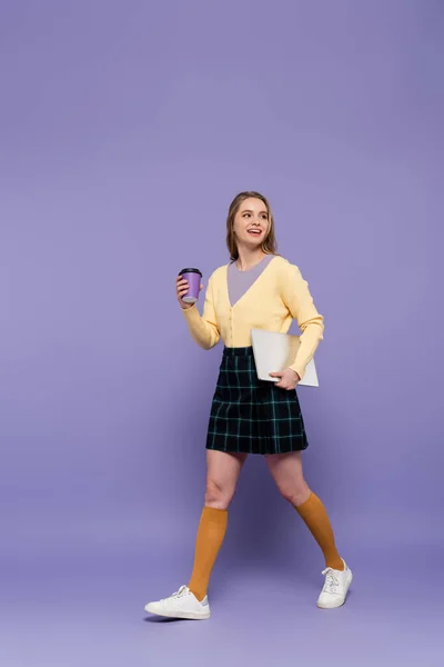 Повна довжина весела молода жінка тримає ноутбук і паперовий стаканчик під час ходьби на фіолетовому — стокове фото