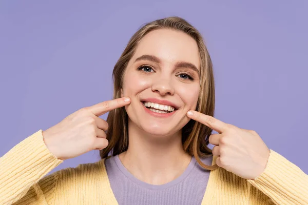 Mujer joven positiva apuntando a las mejillas aisladas en púrpura - foto de stock