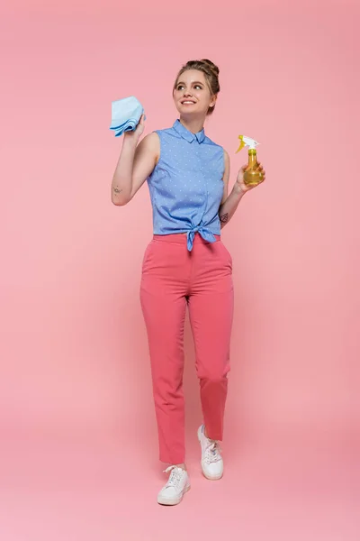 Full length of happy and tattooed woman holding blue rag and spray bottle on pink — Stock Photo