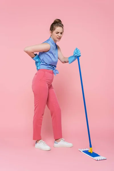 Pleine longueur de jeune femme tatouée dans des gants en caoutchouc tenant la serpillière et touchant le dos sur rose — Photo de stock