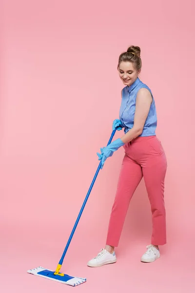 Longitud completa de la joven sonriente en guantes de goma limpieza con fregona en rosa - foto de stock