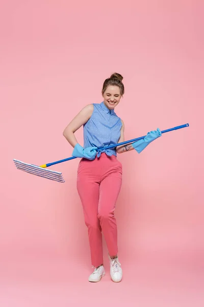 Полная длина веселой молодой женщины в резиновых перчатках имитирует игру на гитаре со шваброй на розовом — стоковое фото