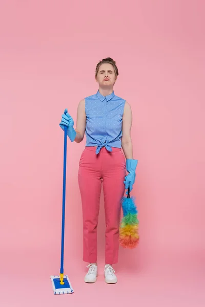 Pleine longueur de bouleversé jeune femme dans des gants en caoutchouc tenant balai et brosse à poussière sur rose — Photo de stock