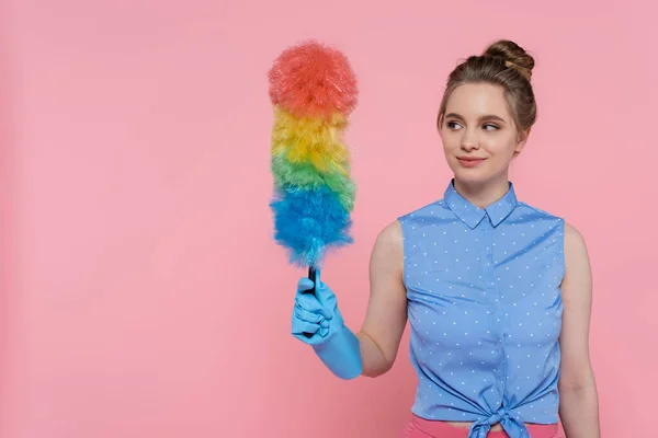 Smiling young woman in blue rubber glove holding dust brush isolated on pink — Stock Photo