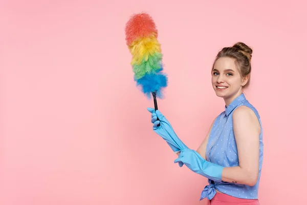 Cheerful and tattooed young woman in blue rubber gloves holding dust brush isolated on pink — Stock Photo