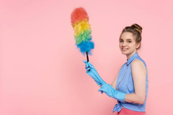 Alegre y tatuada mujer joven en guantes de goma azul con cepillo de polvo aislado en rosa - foto de stock