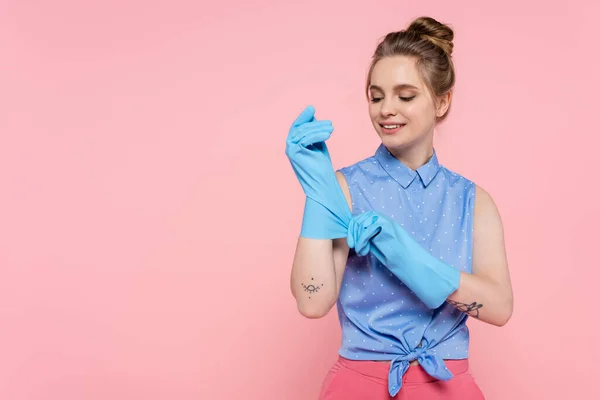 Tätowierte junge Frau trägt blaue Gummihandschuhe, während sie isoliert auf rosa lächelt — Stockfoto