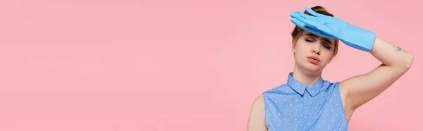 Tired and tattooed young woman in rubber glove wiping sweat isolated on pink, banner — Stock Photo