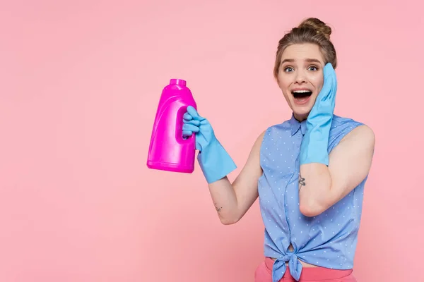Збуджена молода жінка в гумових рукавичках тримає яскраву пляшку з миючим засобом ізольовано на рожевому — стокове фото