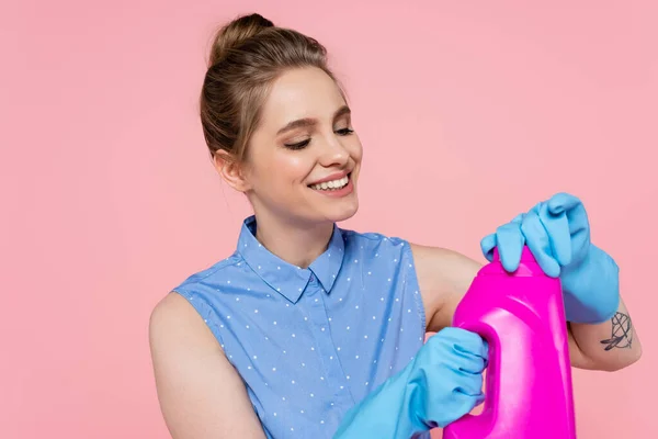 Joven sonriente con guantes de goma sosteniendo botella brillante con detergente aislado en rosa - foto de stock