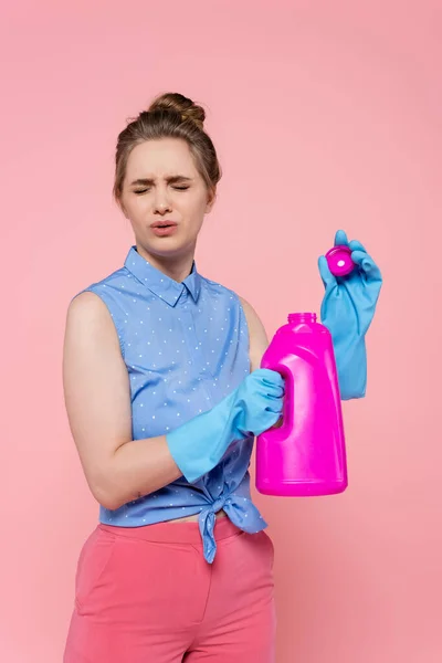 Mulher jovem descontente em luvas de borracha segurando garrafa brilhante com detergente isolado em rosa — Fotografia de Stock
