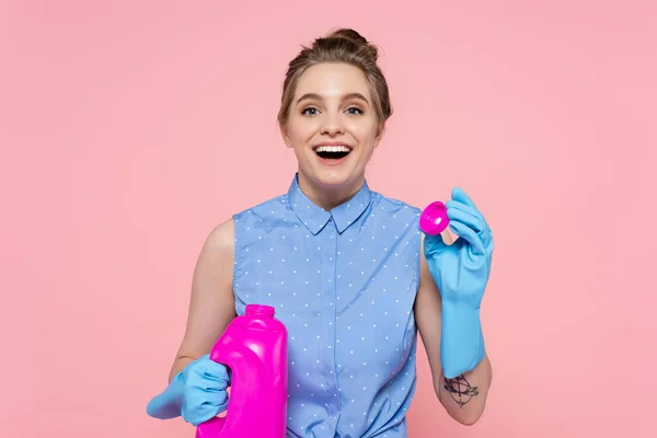 Stupito giovane donna in guanti di gomma tenendo bottiglia luminosa con detergente isolato su rosa — Foto stock