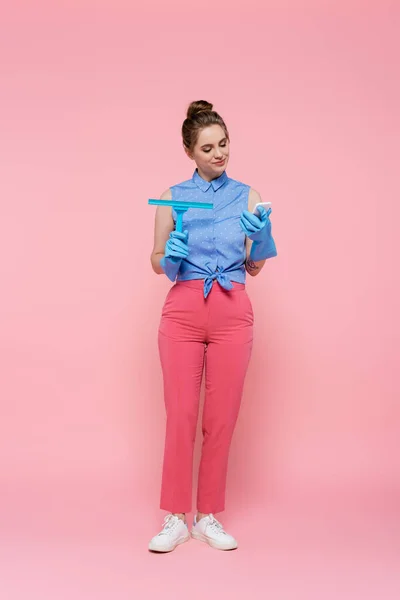 Full length of cheerful young woman in rubber gloves holding window wiper and using smartphone on pink — Stock Photo