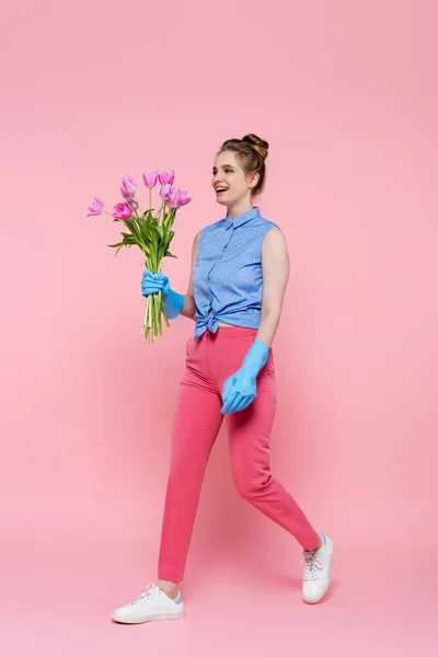 Full length of happy young woman in rubber gloves holding tulips and walking on pink — Stock Photo