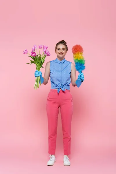 Повна довжина щасливої молодої жінки в гумових рукавичках, що тримає тюльпани і пил щітка на рожевому — стокове фото